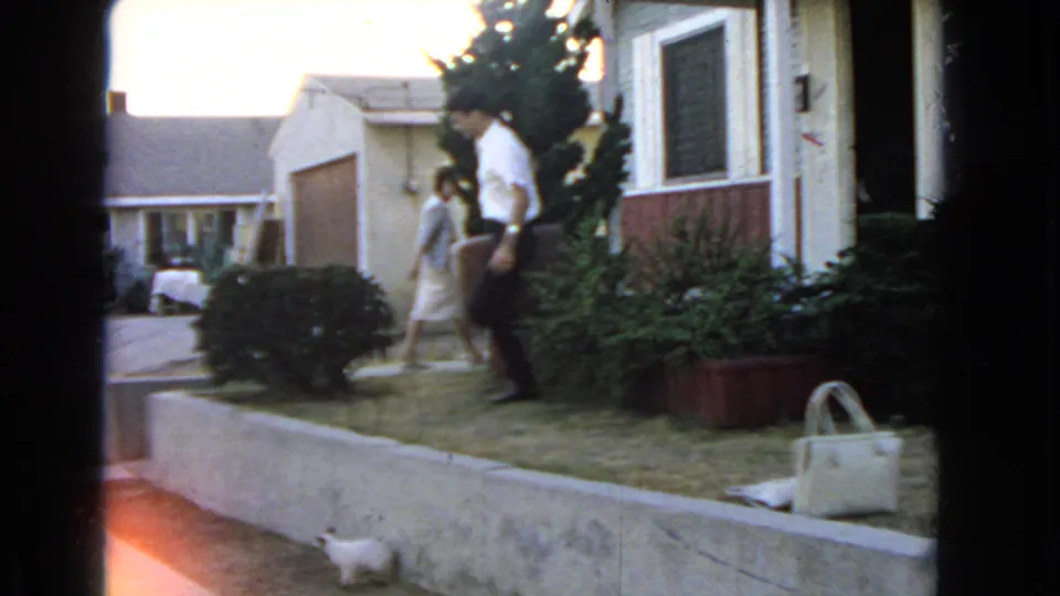 A man in a white shirt walks past a woman on a San Pedro lawn, with a handbag visible near the house.