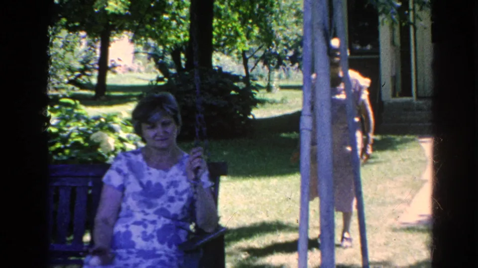 A woman in a blue patterned dress sits on a porch swing as another woman in a light dress stands near the house path.