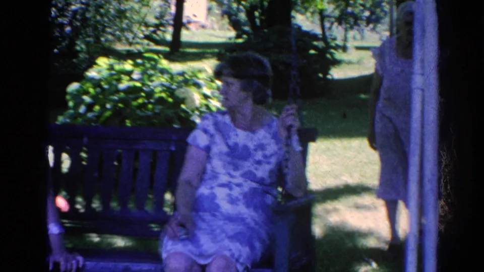A woman in a patterned dress sits on a porch swing in a leafy Silver Lake garden, with another figure partly visible at frame edge.
