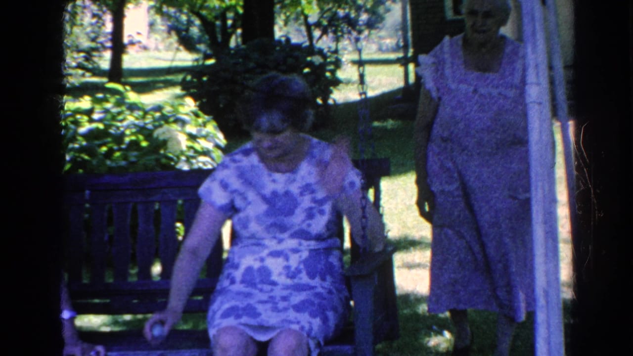 1966 Silver Lake home-movie frame showing an adult woman seated by a wooden garden bench while another woman stands in shade.