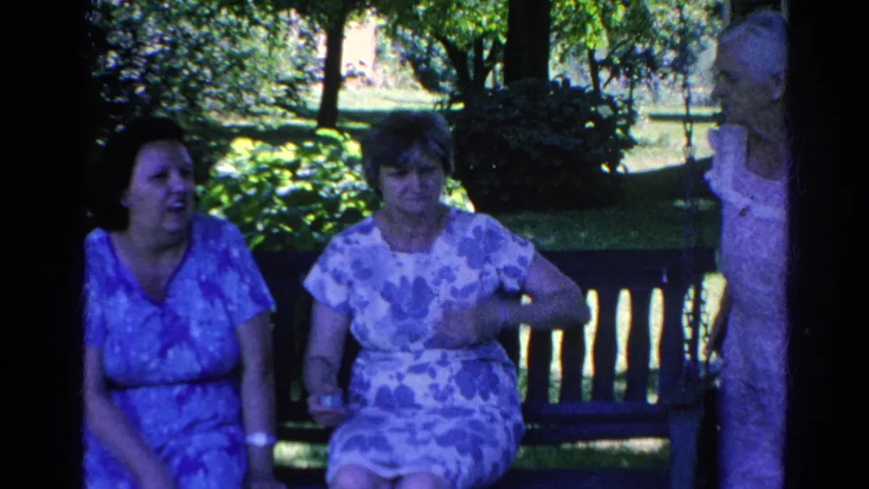 Two adult women sit on a wooden bench in a shaded garden while a third woman stands nearby in a patterned dress.