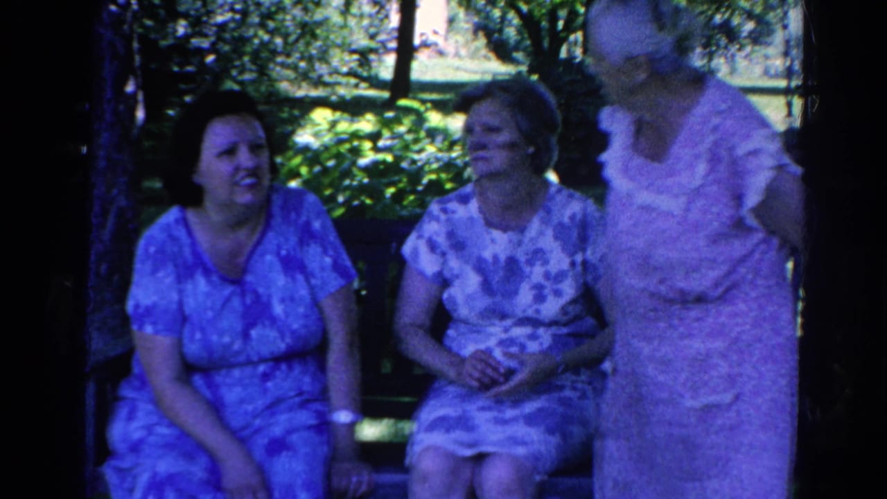 Three adult women in patterned dresses gather beside a park bench outdoors.
