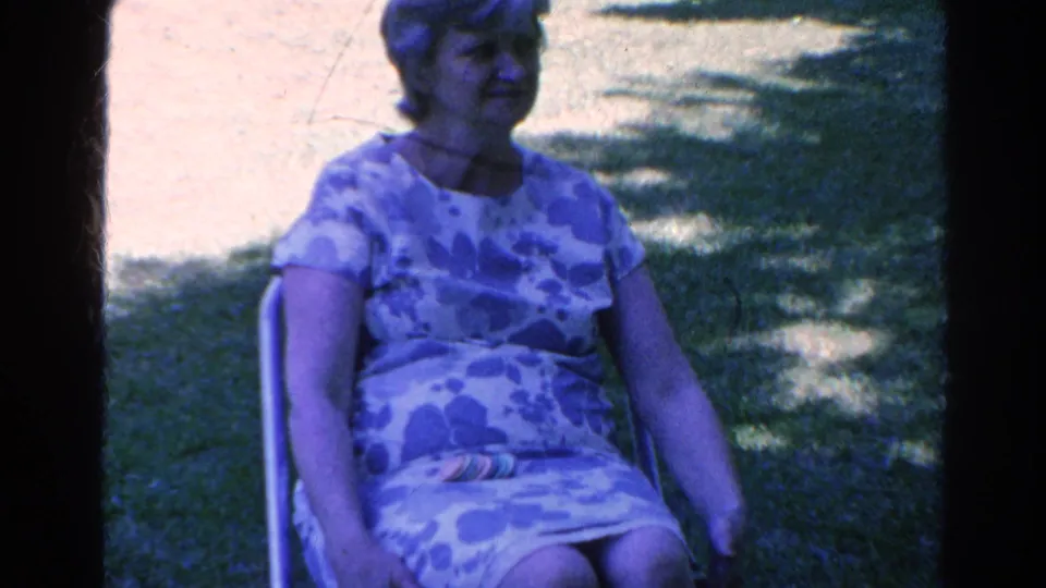 A woman sits alone in a patterned dress in the Silver Lake outdoor home-movie setting, with lawn and tree shade behind her.