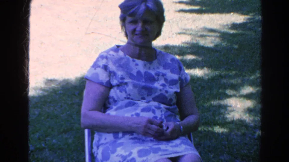 A woman in a patterned dress sits facing the camera in the 1966 Silver Lake home-movie footage, surrounded by lawn and tree shade.