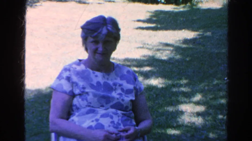 A closer 8mm frame shows the seated woman in a patterned dress, with grass and shifting tree shade behind her.
