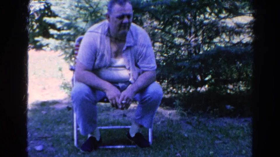 A man in a light shirt and pants sits in a folding chair outdoors, near garden foliage in the same home-movie sequence.