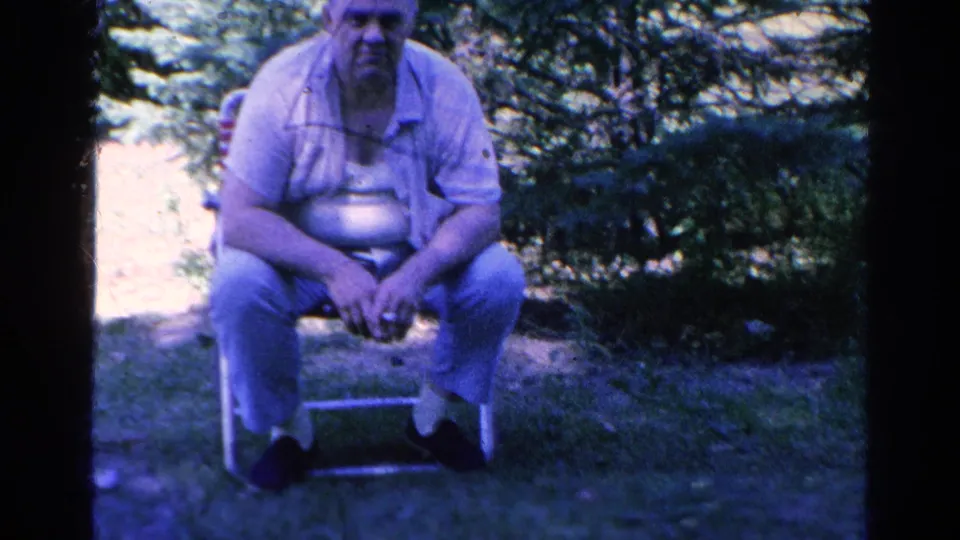 In a 1966 Silver Lake yard scene, a man sits on a folding chair under tree shade with grass and shrubs behind him.