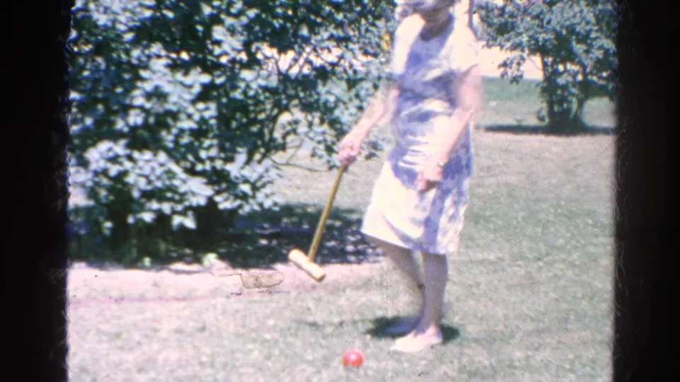 A woman in a light patterned dress stands in the 1966 Silver Lake yard with a wooden croquet mallet near a red ball.