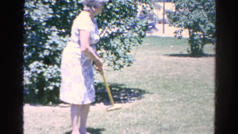 The woman turns across the Silver Lake backyard lawn holding a croquet mallet, with shrubs and trees framing the grass.