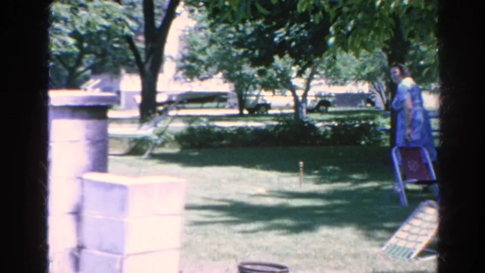 A woman stands near a folding chair on the Silver Lake lawn, with trees, stone pillars, and shaded grass behind her.