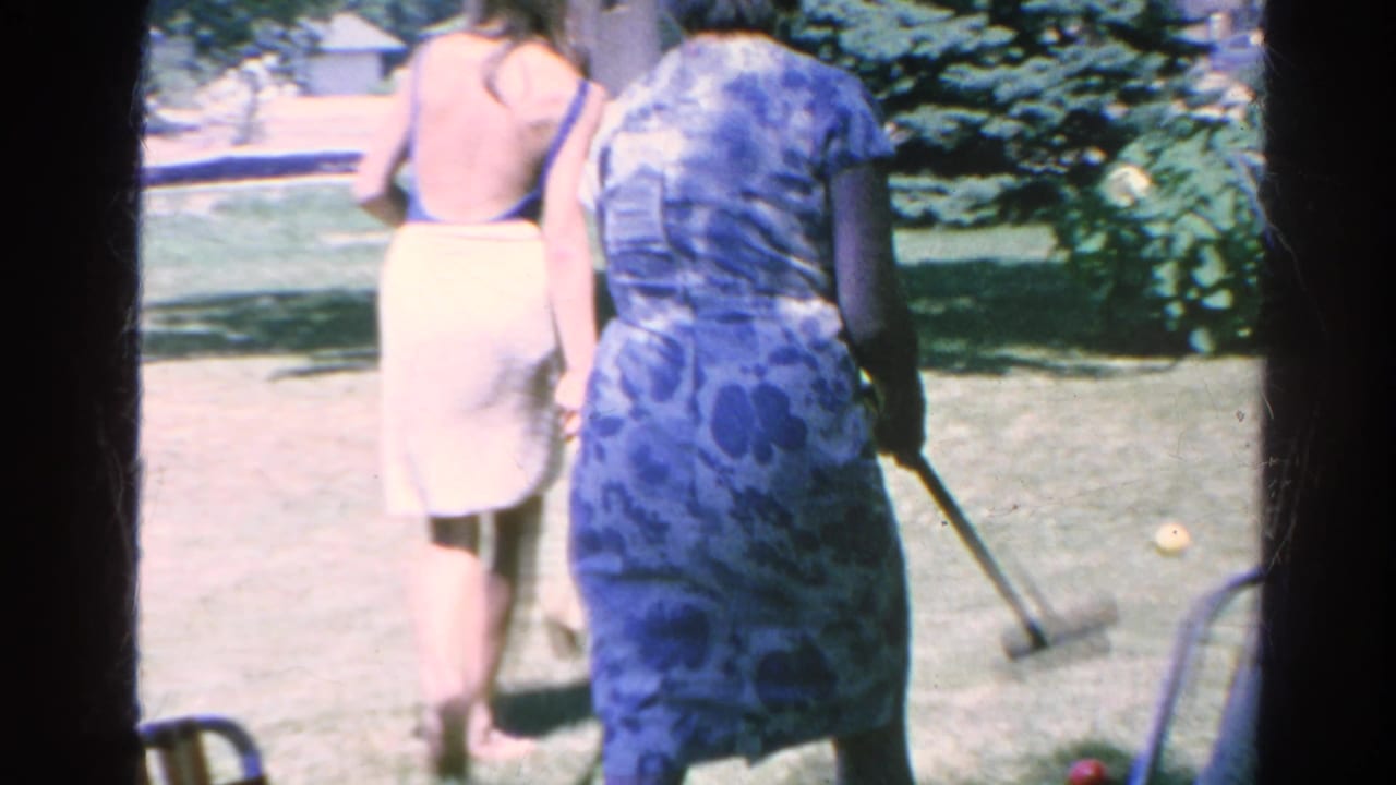 Two women walk across a lawn, one holding a croquet mallet.