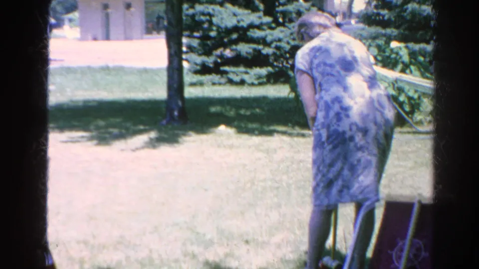 A woman in a blue patterned dress bends toward the grass during a 1966 Silver Lake backyard croquet game.