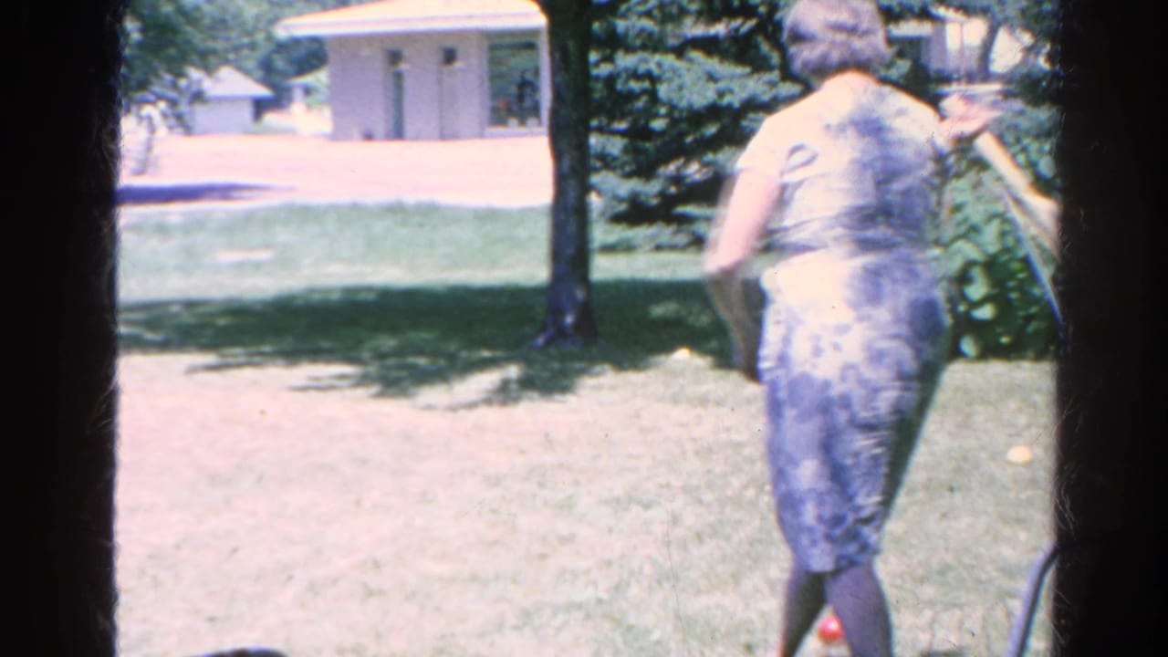 Woman seen from behind holding a croquet mallet on a sunny lawn near a hammock.