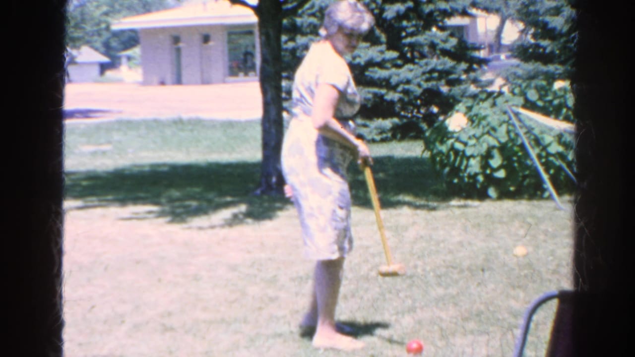 Woman stands with a croquet mallet beside a red ball on grass, with a hammock behind her.