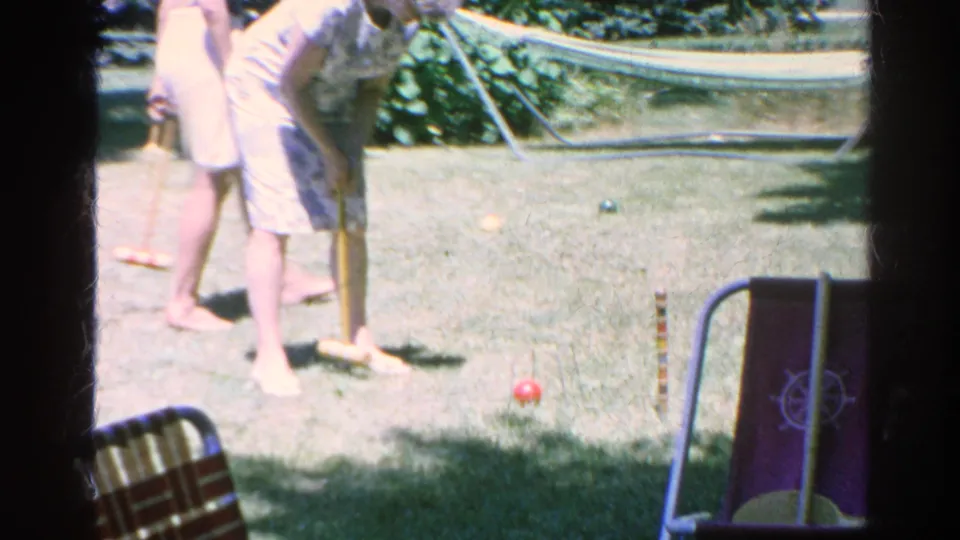 A barefoot woman swings a croquet mallet near wickets and colored balls in a sunlit 1966 Silver Lake backyard home movie.