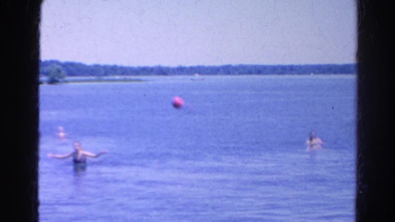 Swimmers in shallow lake water with a red ball and distant shoreline.