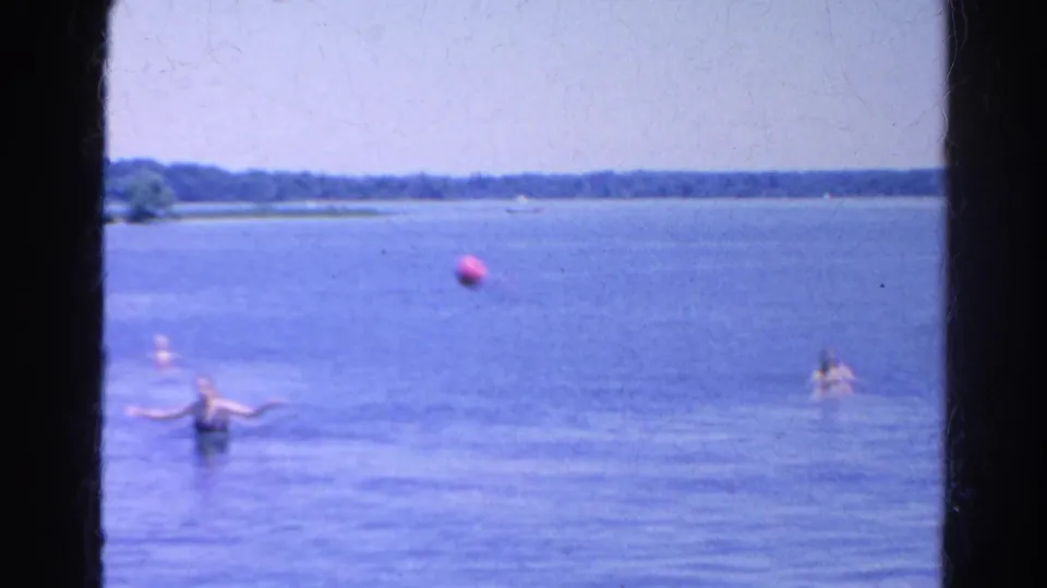1966 Silver Lake home-movie frame showing swimmers in shallow blue water as a red ball floats nearby.