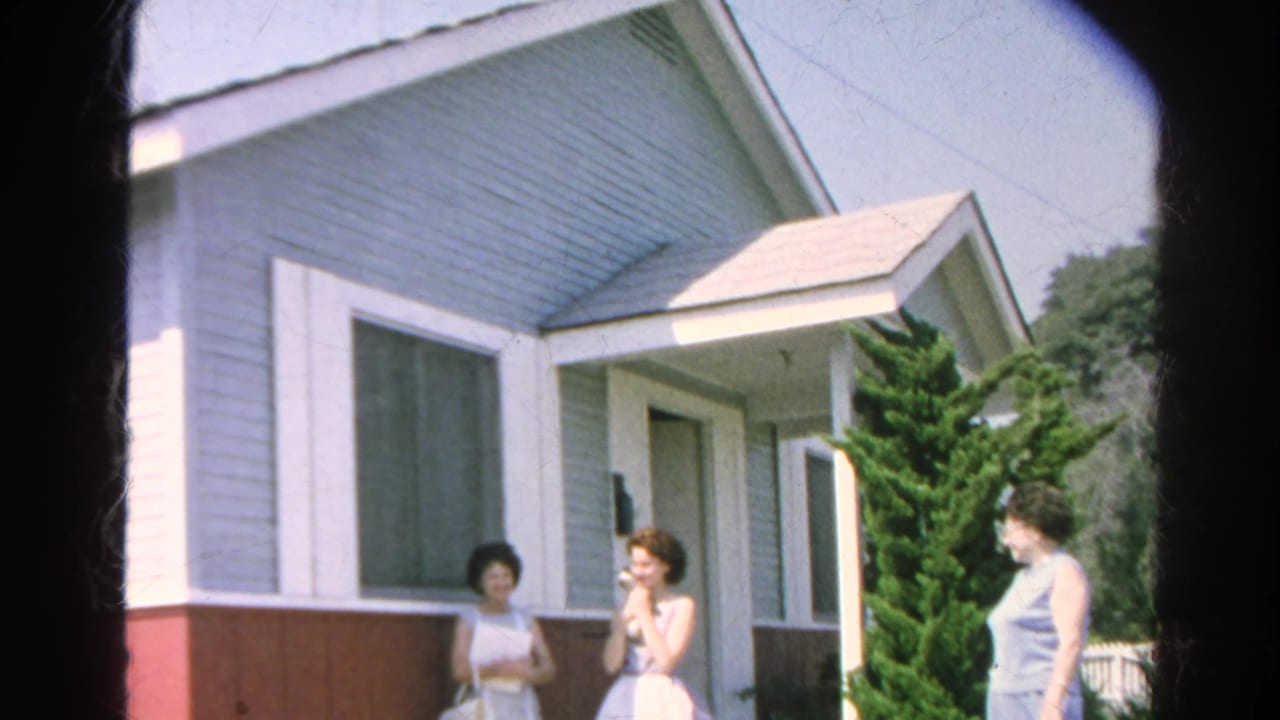 1966 Silver Lake home-movie frame showing three adult women outside a light blue house with red lower siding and shrubs.