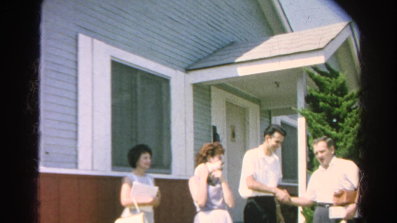 Adults stand outside a light blue house as a young man shakes hands with an older man.