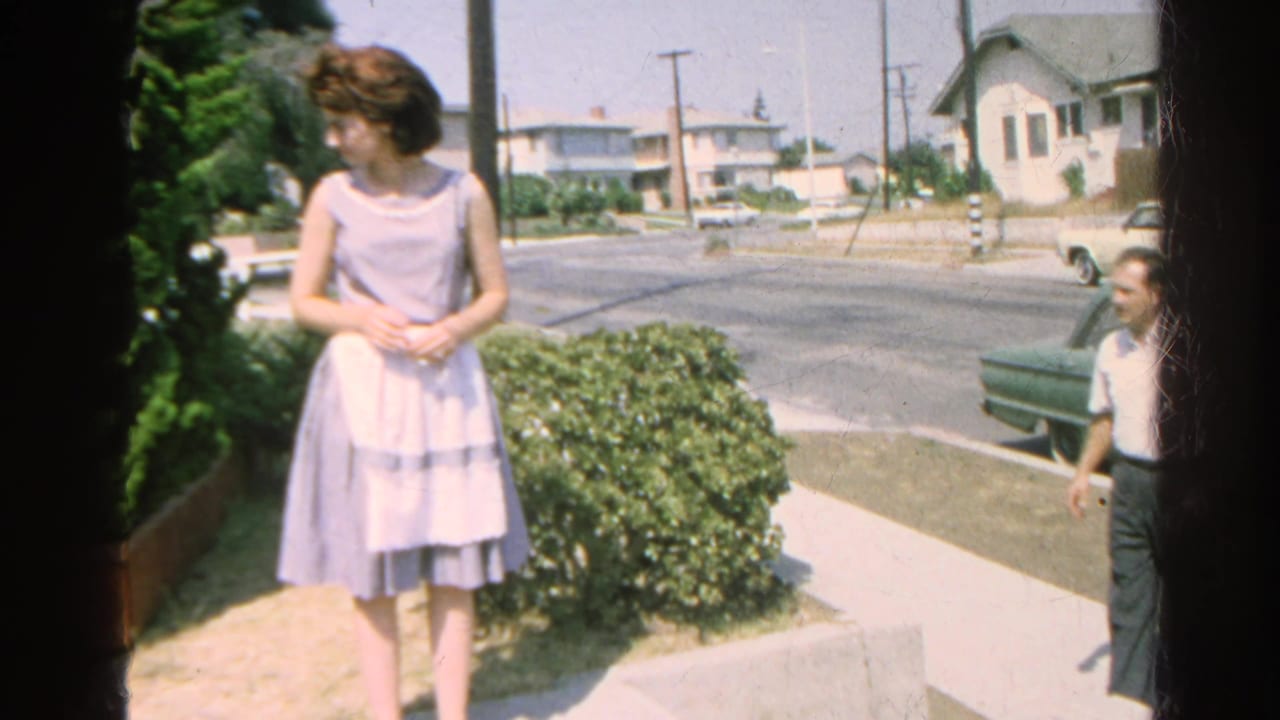 Woman in a light dress stands by shrubs as a man approaches near a street.