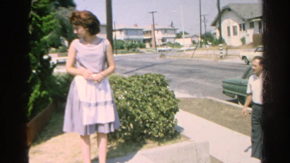 In Silver Lake, a woman in a light 1960s dress stands by shrubs while a man approaches near a residential street and parked car.