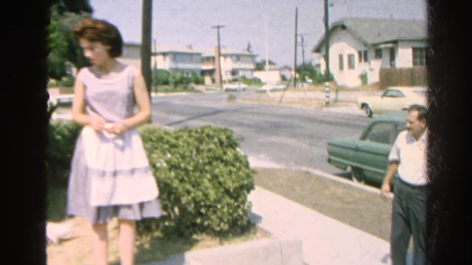 A woman in a light dress stands beside shrubs as a man looks down toward a white cat near the edge of a Silver Lake sidewalk.