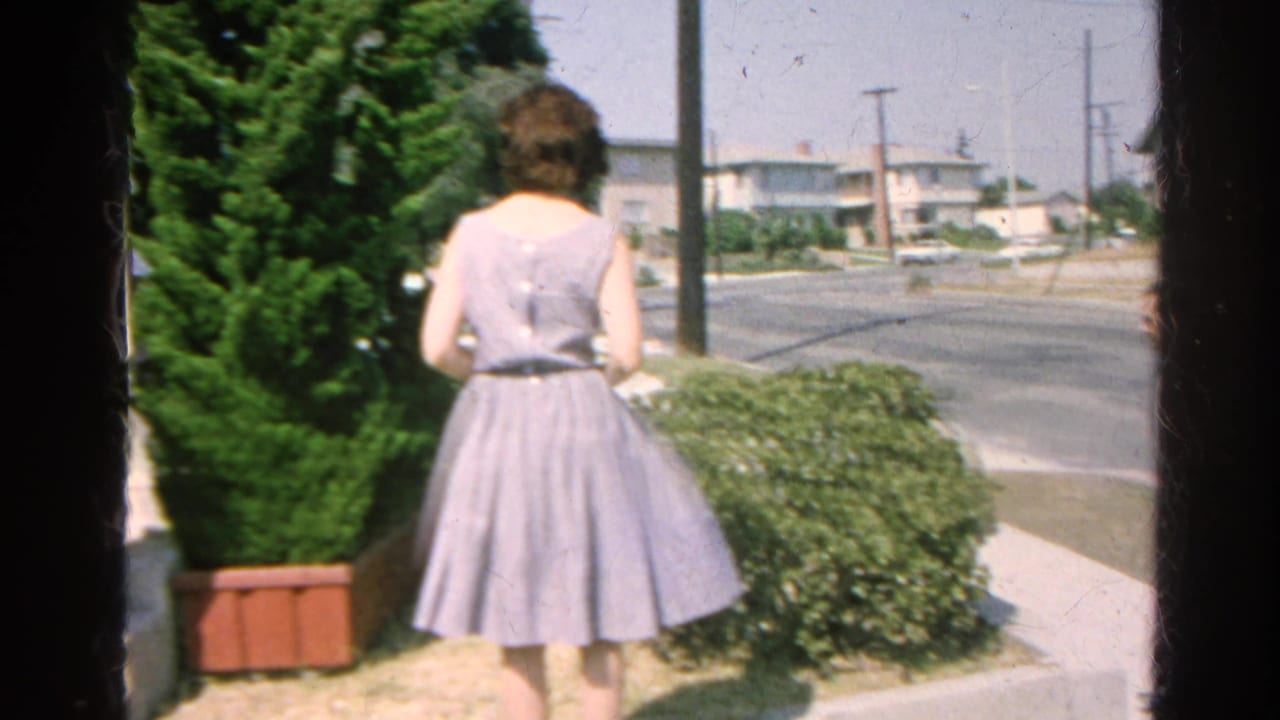 Woman seen from behind in a light dress near shrubs and a residential street.