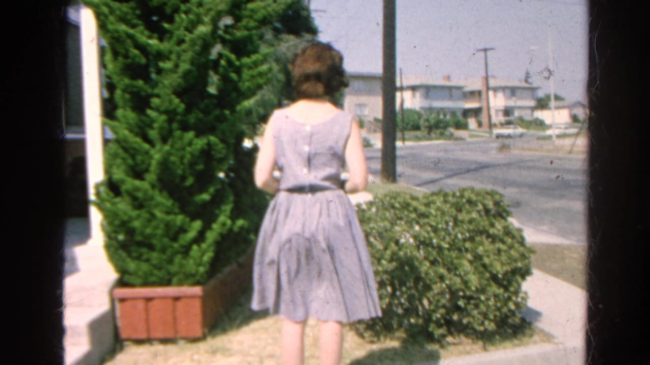Woman in a light dress stands near a trimmed shrub and street, viewed from behind.
