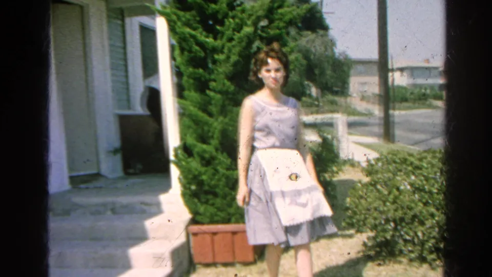 A woman in a light tea-length dress faces the camera and smiles near a house entrance, shrubs, and a potted evergreen in Silver Lake, California, 1966.