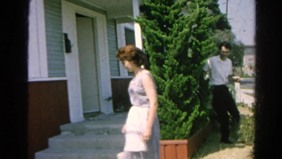 The woman stands beside the house and shrubbery as a man in a white shirt walks behind her near the sidewalk in 1966 Silver Lake home-movie footage.