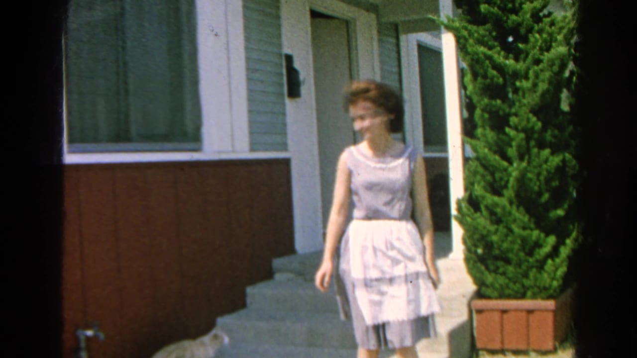 Woman in a light dress stands on house steps beside an open door and tall shrub.