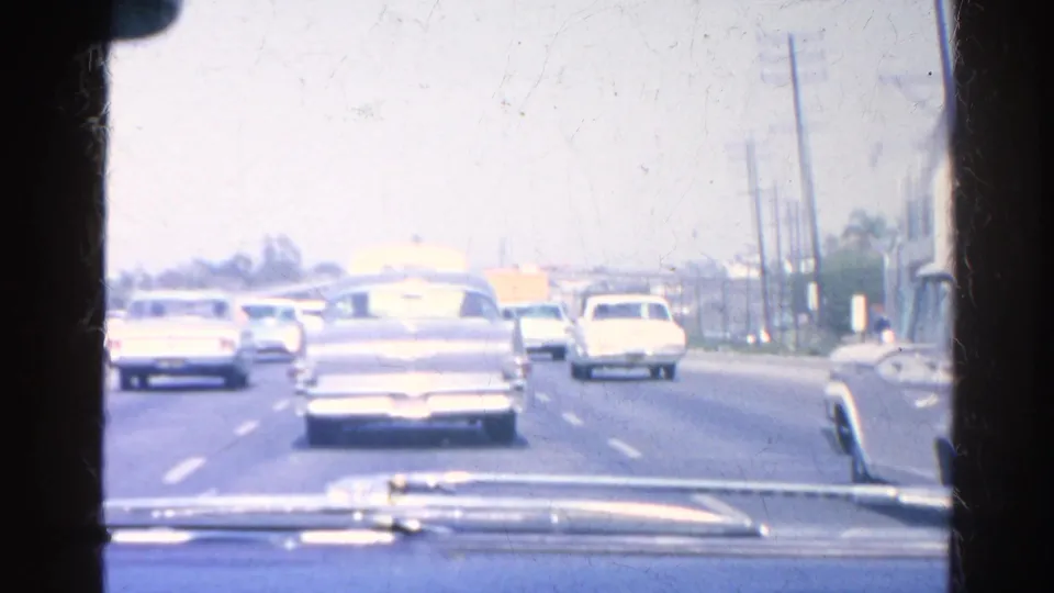 A windshield view shows classic American cars traveling on a bright Southern California highway in the 1966 home-movie sequence.