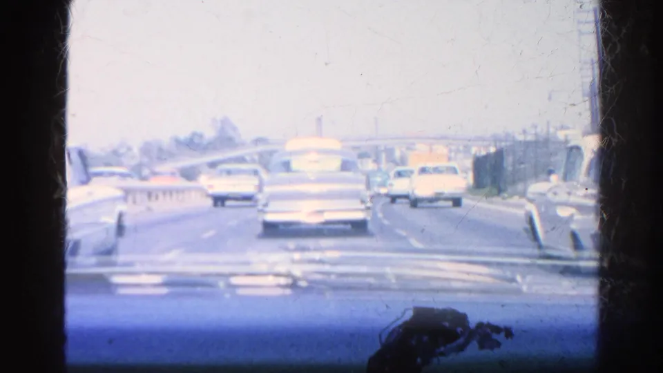 Final in-car frame from the 1966 Silver Lake clip, showing 1960s cars ahead, a roadway barrier or divider, roadside poles, and a blue dashboard foreground.