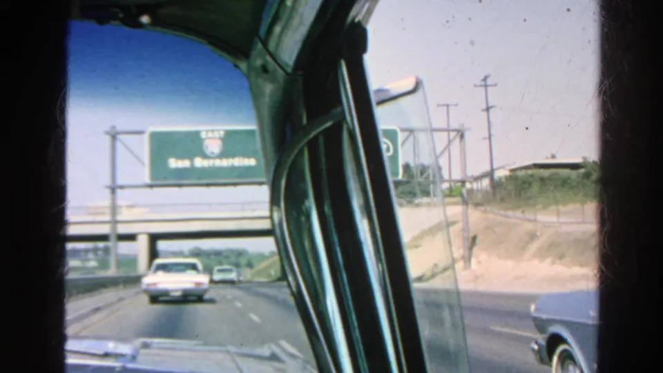 1966 Silver Lake-area freeway drive seen through a passenger-side car window, with classic cars and a San Bernardino guide sign ahead.