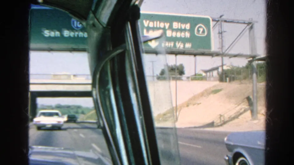 1966 freeway footage from inside a vintage car, showing overhead signs for San Bernardino and Valley Blvd with vehicles ahead.
