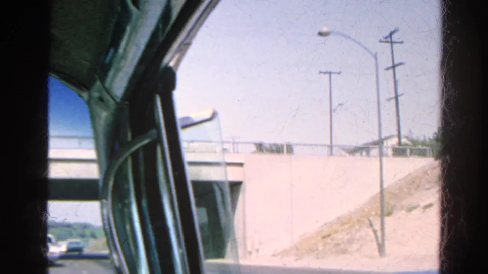 Passenger-side 8mm view during a 1966 freeway drive, with an overpass, utility poles, and dry roadside embankment visible.