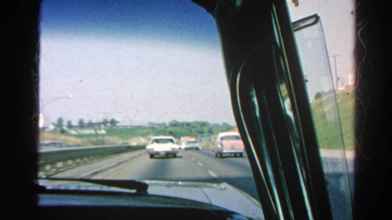 Cars travel ahead on a sunny freeway, framed by the car windshield and side window.