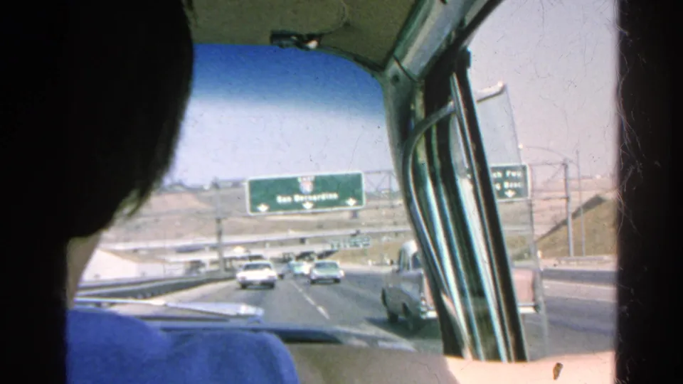 Passenger-side view from a 1966 Southern California freeway drive, with overhead guide signs and midcentury cars ahead.