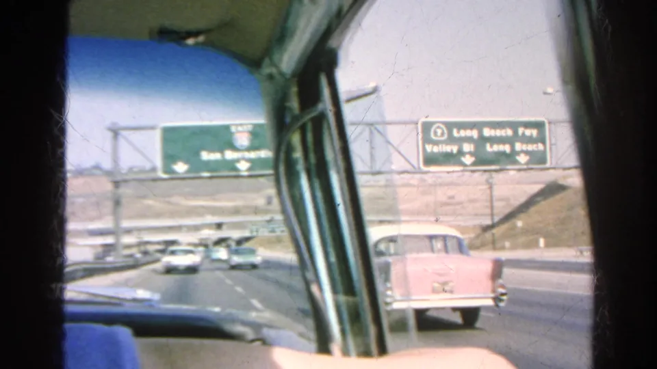 1966 freeway traffic near Silver Lake with a pink midcentury car, overhead route signs, and the view framed by a car window.