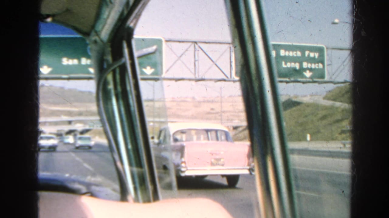 1966 Southern California car-window view showing freeway traffic, a pale wagon ahead, and green signs for Long Beach routes.