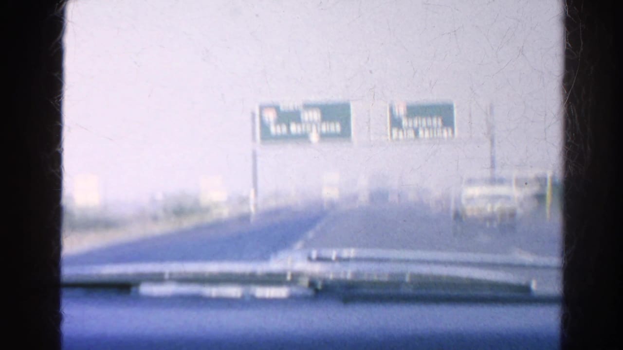 Car-mounted 8mm view of overhead freeway guide signs for San Bernardino and Palm Springs with a vehicle ahead on the road.