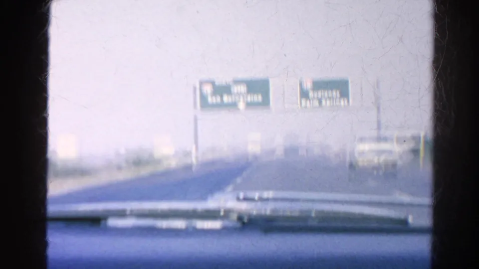 Car-mounted 8mm view of overhead freeway guide signs for San Bernardino and Palm Springs with a vehicle ahead on the road.