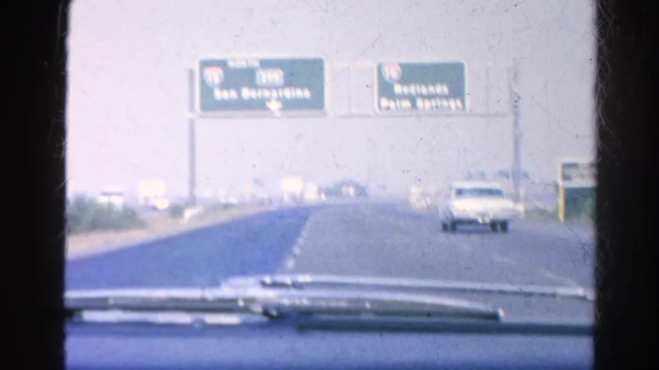 1966 home-movie view through a windshield toward overhead signs for San Bernardino and Redlands or Palm Springs, with a car approaching in the next lane.