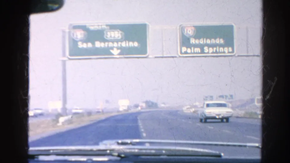 1966 8mm windshield view of overhead Interstate guide signs for San Bernardino, Redlands, and Palm Springs with light freeway traffic ahead.
