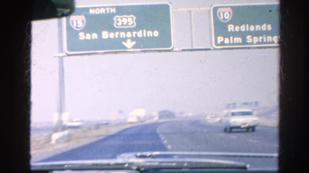View through a car windshield toward overhead freeway signs and traffic on a hazy road.