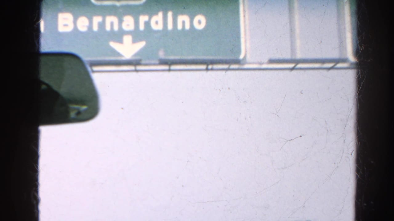 Interior car view beneath a green San Bernardino freeway sign, with the rearview mirror and windshield edge visible.