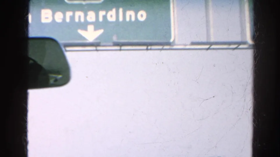 Interior car view beneath a green San Bernardino freeway sign, with the rearview mirror and windshield edge visible.