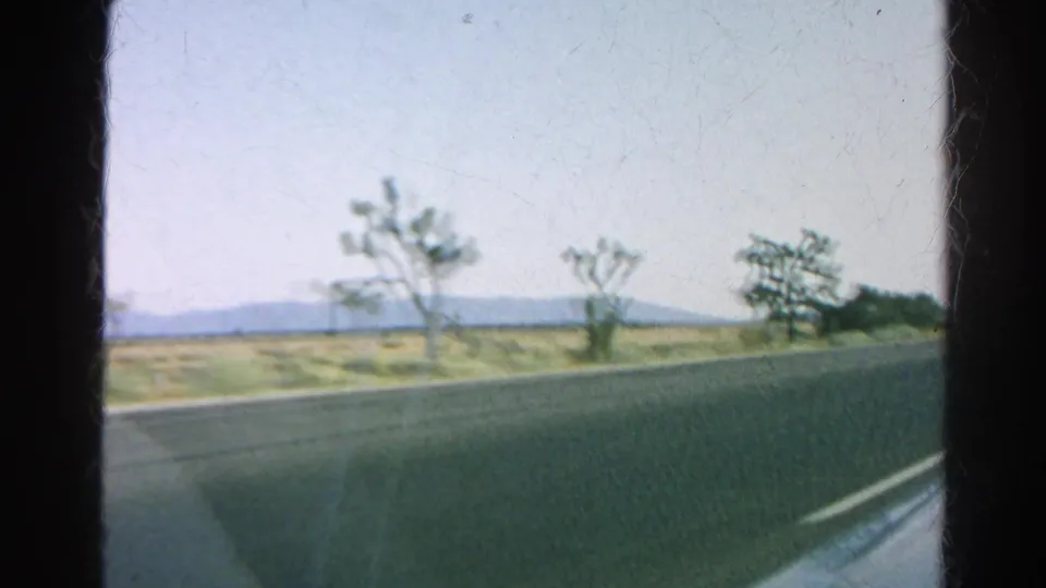 1966 8mm side-window view of desert roadside, Joshua trees, distant hills, and highway pavement in California.