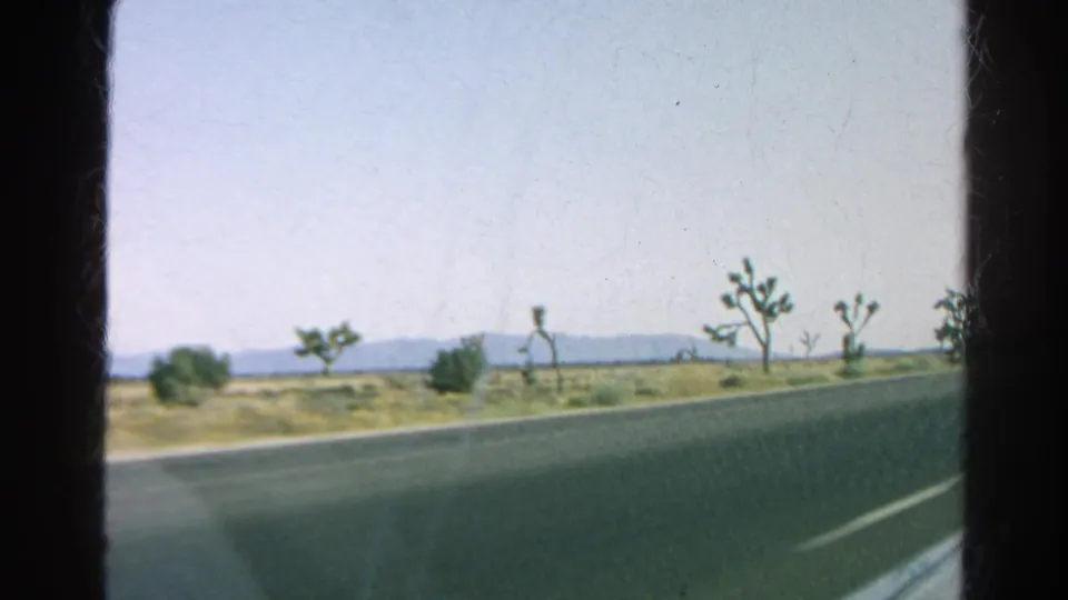 Side-window road-trip view of Joshua trees, scrubland, and distant mountains beside a California highway.