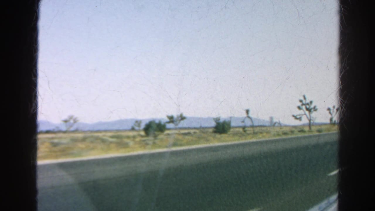 Paved roadside passing a dry desert landscape with shrubs, Joshua trees, distant mountains, and dark frame edges.